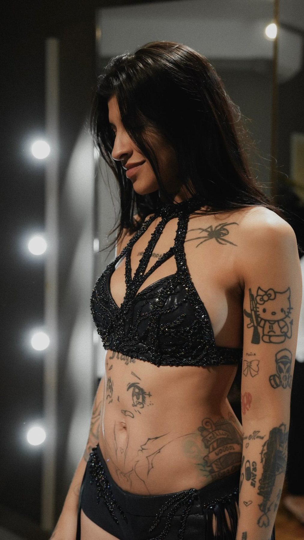 Cazzu backstage in a beaded black harness top, smiling down beside a lit vanity mirror