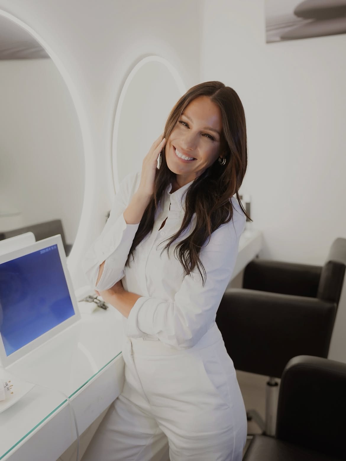 Pampita in a white shirt, hand at her cheek, smiling beside a round lit mirror