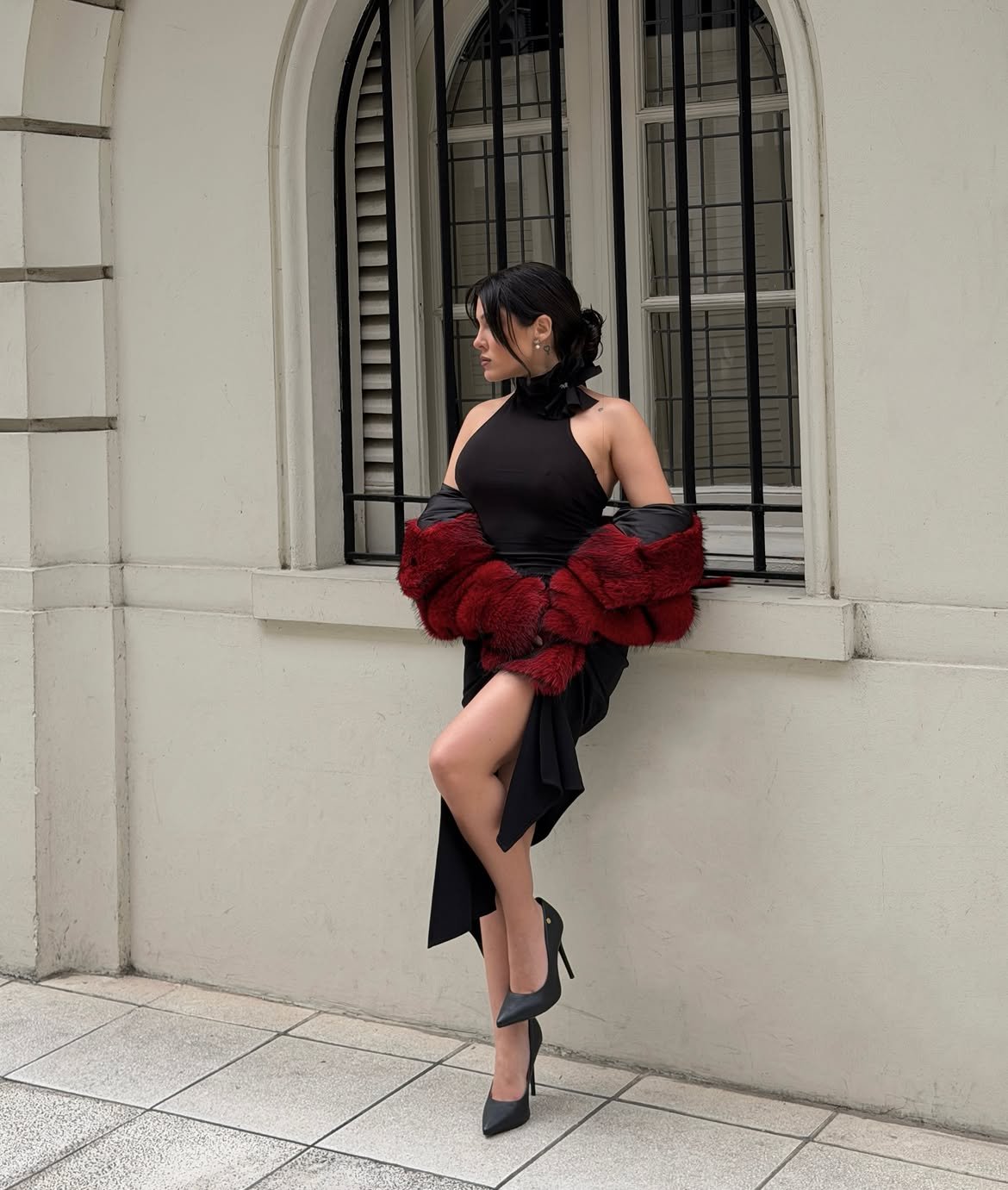 Model in a black halter dress with a red fur stole at an arched window