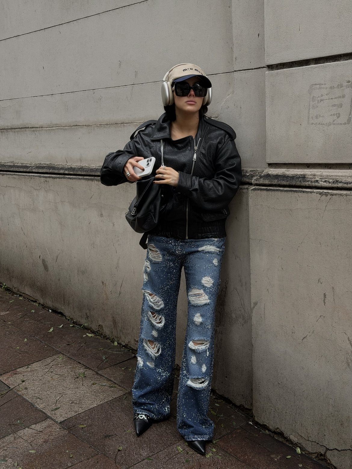 Black leather bomber, crystal-studded ripped jeans, Miu Miu cap and headphones against a grey stone wall