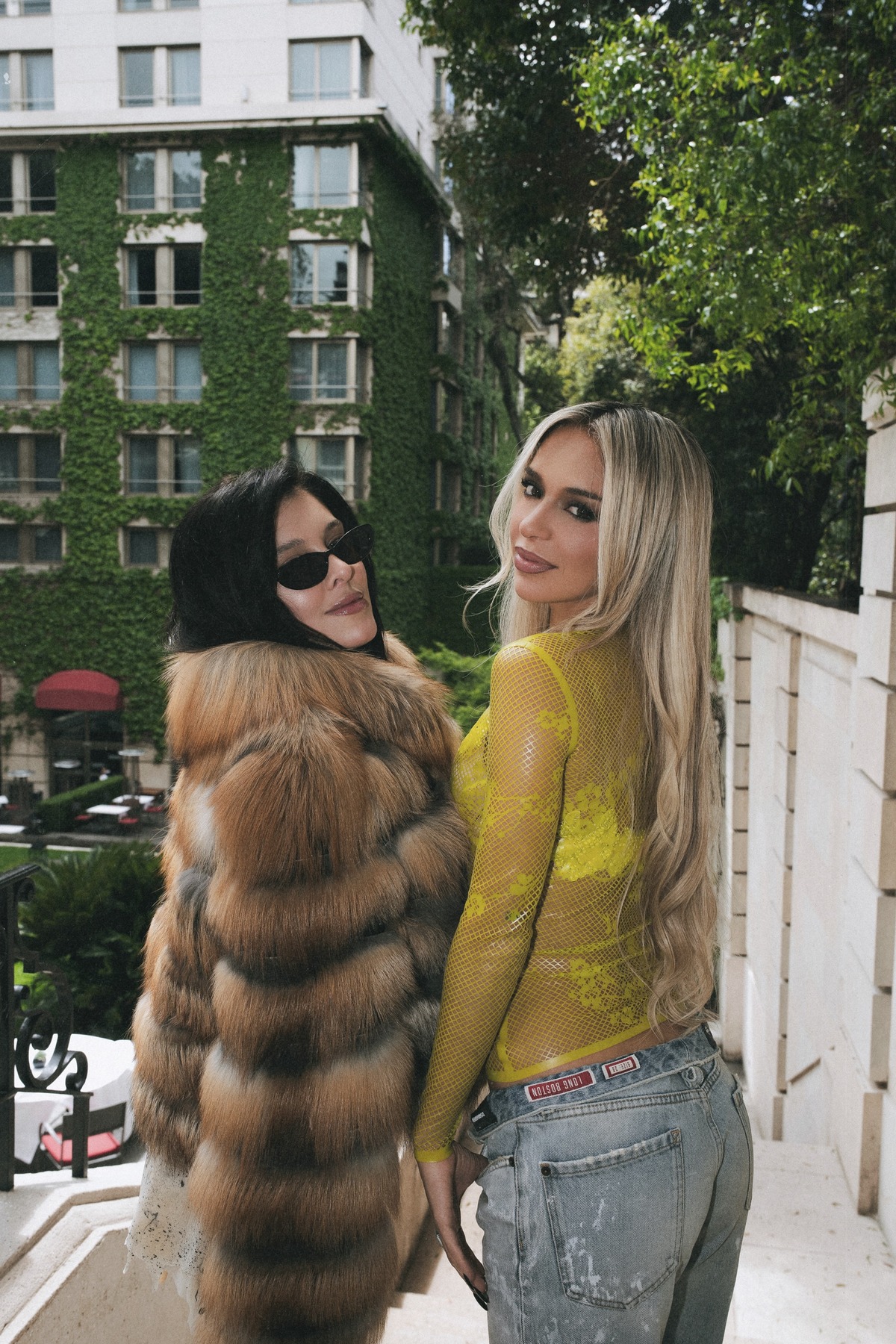 Sofía Gonet and Bad Gyal glancing back on a hotel terrace above an ivy-covered façade